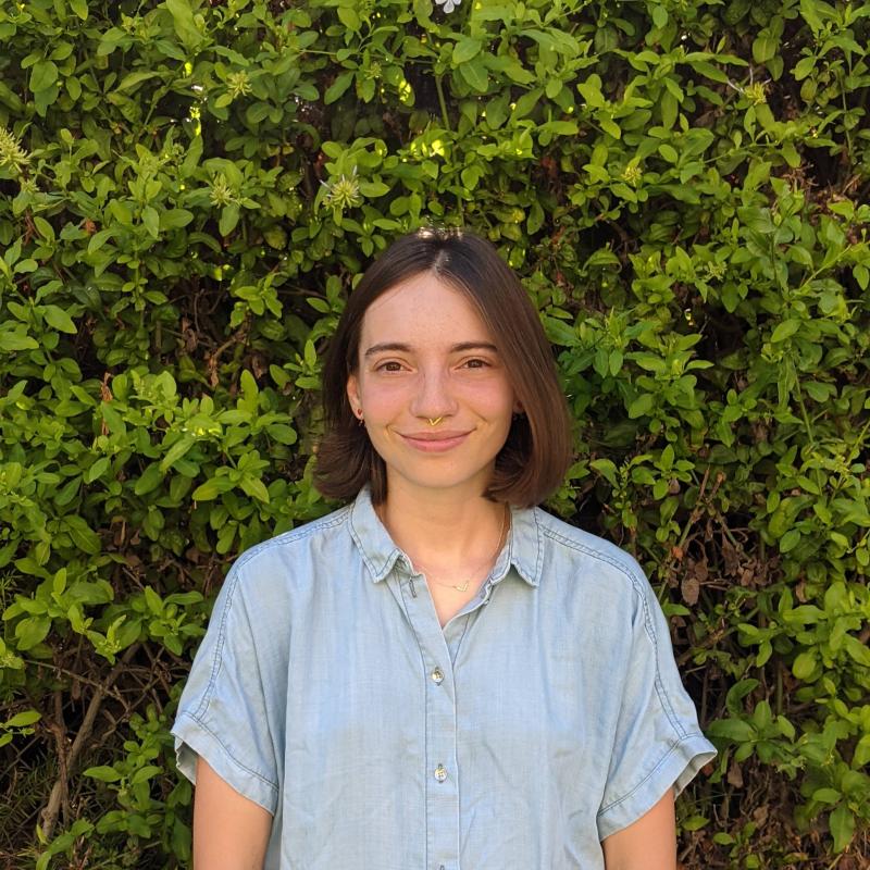 Karina is wearing a blue button up shirt. They are standing in front of a green bush.